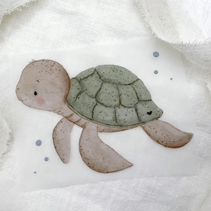Puede incluir: Una ilustración en acuarela de una tortuga marina con cabeza y cuerpo de color marrón claro moteado, con un caparazón verde salvia con contornos oscuros. La tortuga está rodeada de pequeñas burbujas azules y se ve un diminuto corazón negro.