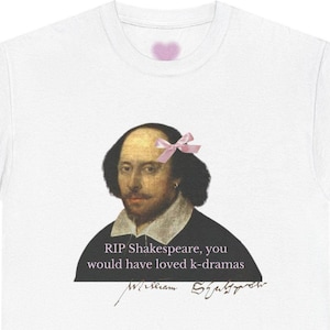 Può includere: T-shirt bianca con un ritratto di William Shakespeare con un fiocco rosa. Il testo recita "RIP Shakespeare, you would have loved k-dramas" con la sua firma. Un cuore rosa è sopra il ritratto.