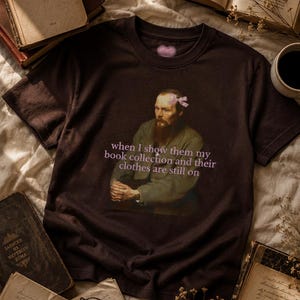 Può includere: Una t-shirt a maniche corte marrone scuro con la grafica di un uomo barbuto che indossa un piccolo fiocco rosa sulla testa. Il testo "when I show them my book collection and their clothes are still on" è stampato in lettere rosa chiaro.