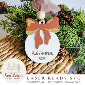 May include: A white, round Christmas ornament with a bow design in shades of orange and gold. The ornament has the name "niemann" and the year "2025" engraved on it. It is displayed on a woven surface with greenery and pinecones.