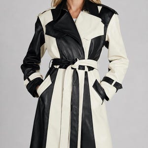 May include: A long, two-tone trench coat in black and cream. The coat features a belted waist, notched lapels, and a geometric colour-block design. The sleeves have black cuffs, and the coat has pockets.