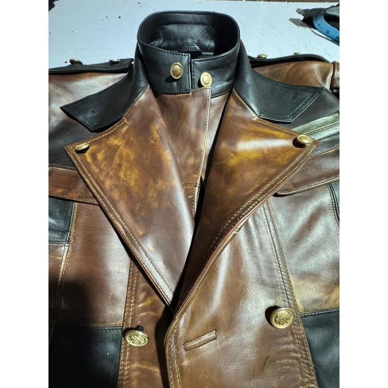 May include: A brown and black leather jacket with a high collar and gold buttons. The jacket has a double-breasted design and a distressed finish, giving it a vintage look. The leather appears to be high-quality and well-crafted.