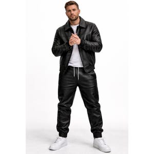 Puede incluir: Una chaqueta de cuero negra con cuello y pantalones de cuero negro a juego. Los pantalones tienen cintura con cord&oacute;n y pu&ntilde;os el&aacute;sticos. El conjunto se combina con zapatillas blancas. El modelo est&aacute; de pie frente a un fondo blanco.