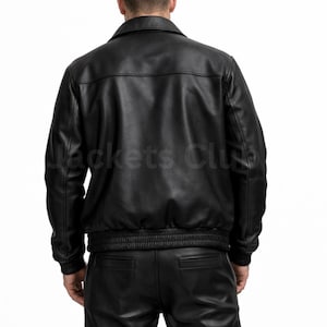 Puede incluir: Chaqueta de cuero negra con cuello, cintura y pu&ntilde;os el&aacute;sticos. La parte trasera de la chaqueta es visible, con el texto "Jackets Club" impreso en la parte inferior izquierda. La chaqueta es parte de un conjunto de ropa de cuero.