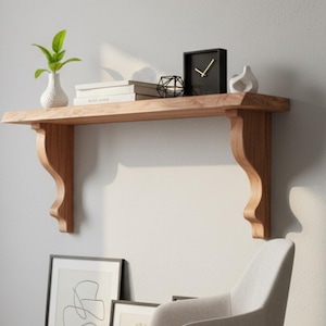 May include: A wooden floating shelf with decorative corbels, holding a small white vase with greenery, books, and a black clock. Below, a framed abstract art print and a beige armchair sit on a gray rug.