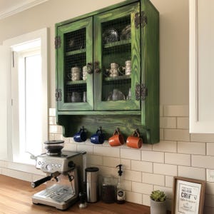 Può includere: Un mobile in legno verde con ante a rete metallica, che espone stoviglie. Sotto, quattro tazze sono appese a ganci. Una macchina per l'espresso in acciaio inossidabile è sul bancone, insieme ad accessori per il caffè.