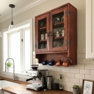 Peut inclure: Un meuble de cuisine mural en bois brun foncé avec des portes vitrées et des inserts en treillis métallique. Le meuble est orné de ferrures décoratives en métal. Quatre tasses sont suspendues en dessous. Ambiance cuisine.