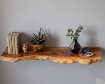 May include: A live-edge wooden shelf displays a small succulent in a wooden bowl, a stack of books, a ceramic owl figurine, a black geometric vase with greenery, and decorative stones. The shelf is mounted on a gray wall.