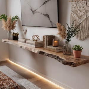 May include: A live-edge wooden shelf mounted on a white wall, adorned with decorative items. The shelf holds books, vases with dried plants, a geometric gold object, and a small potted succulent. A macrame wall hanging and a painting are also visible.
