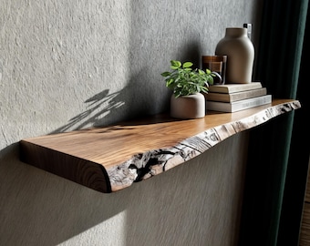 May include: A wooden floating shelf with a natural edge, in a warm brown hue. The shelf is mounted on a grey wall and displays a small potted plant, books, and a vase. The shelf's unique edge adds a rustic touch to the decor.
