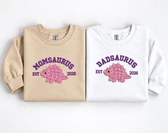 Könnte beinhalten: Ein beigefarbener Pullover und ein weißer Pullover, jeweils mit einer gestickten rosa Ankylosaurus-Grafik. Das beige Oberteil zeigt den Text MOMSAURUS EST 2026, während das weiße Oberteil den Text DADSAURUS EST 2026 in violetter Schrift zeigt.