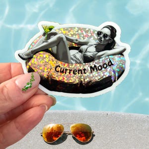 May include: A sticker with the text "Current Mood" features a grayscale illustration of a person relaxing in a pool float with a cocktail. The sticker has a glittery border. A pair of gold aviator sunglasses rests below.