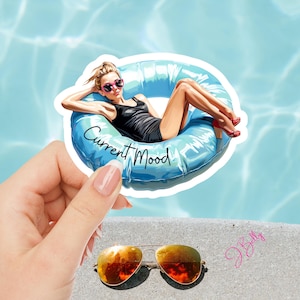 May include: A sticker featuring a woman in a black swimsuit lounging on a blue inflatable ring in a pool. The sticker has the text "Current Mood" and is held near a pair of gold-rimmed sunglasses.