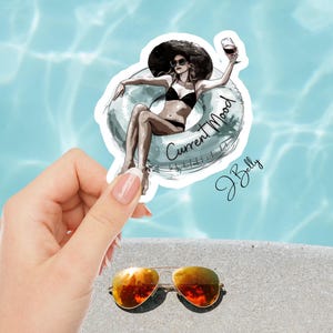 May include: A sticker featuring a woman in a black bikini and a large hat, lounging in a pool float with a glass of wine. The sticker has the words "Current Mood" and the signature "J. Belly". Gold aviator sunglasses are in the foreground.