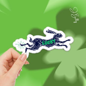 May include: A sticker featuring a running rabbit in dark blue with the word "LUCKY" in green. The sticker has a white border and is held against a green background with a clover design.