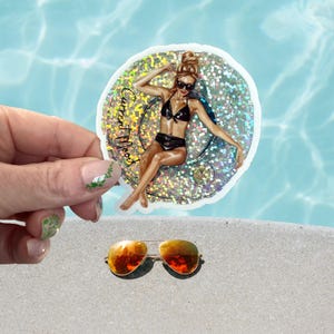 May include: A sticker featuring a woman in a black bikini, sunglasses, and a bun, with the text "Current Mood." The sticker has a glittery, iridescent border. Below, a pair of aviator sunglasses with orange lenses rests on a sandy surface.