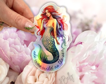 May include: Holographic mermaid sticker with red, orange, and purple hair, a green and blue tail, and a white dress. The sticker has the text "she was wild as the sea." The sticker is held in front of a background of pink flowers.
