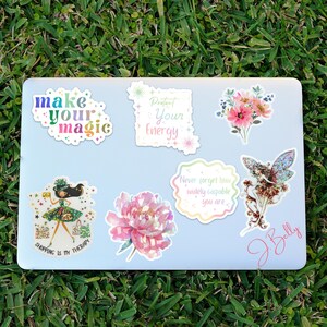 May include: A collection of colorful stickers on a light blue laptop. The stickers feature floral designs, inspirational quotes like "Make Your Magic" and "Protect Your Energy", and a whimsical illustration of a fairy and a woman with shopping bags. The laptop is on a bed of green grass.