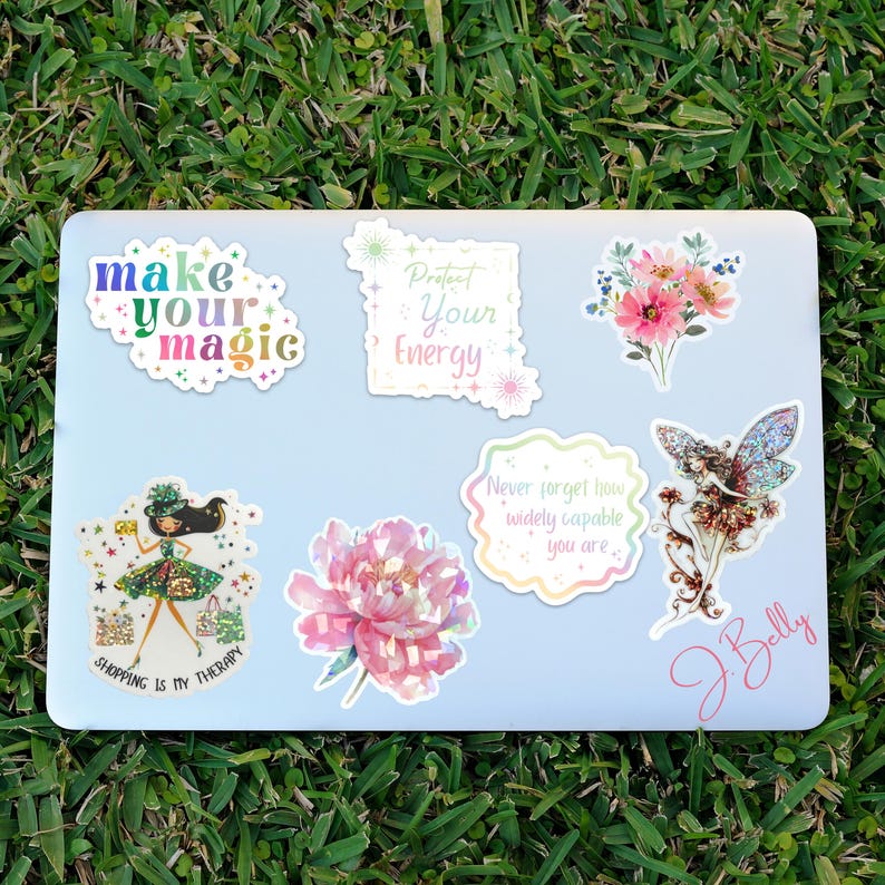 May include: A collection of colorful stickers on a light blue laptop. The stickers feature floral designs, inspirational quotes like "Make Your Magic" and "Protect Your Energy", and a whimsical illustration of a fairy and a woman with shopping bags. The laptop is on a bed of green grass.