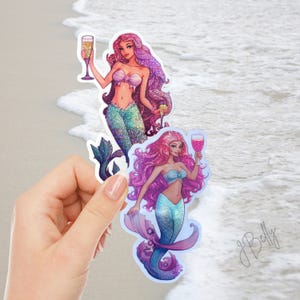 May include: Two die-cut stickers featuring mermaids with long, glittery pink and purple hair. Each mermaid wears a light blue or purple seashell top and has a shimmering green or blue tail. They are each holding a glass of pink champagne.
