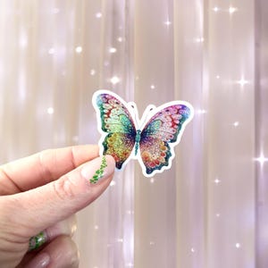 May include: A colorful butterfly sticker with glitter accents. The sticker features a vibrant design with shades of green, blue, red, and gold. The butterfly has a white border and is held by a hand.