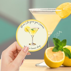 May include: A round yellow coaster with the text "When Life gives you Lemons, make a lemon drop" and a lemon drop cocktail illustration. A hand holds the coaster near a cocktail glass with a lemon slice.