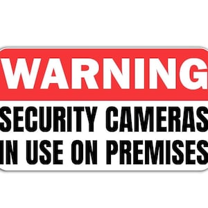 May include: A white rectangular sign with a red band across the top. The red band has the word "WARNING" in white letters. The rest of the sign has the text "SECURITY CAMERAS IN USE ON PREMISES" in black letters.