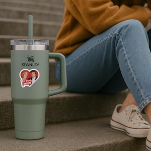 May include: A sage green Stanley tumbler with a handle, a clear lid, and a matching straw. A heart-shaped sticker is applied to the front, featuring two men and the text Best Friends Forever in white script against a red background.