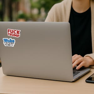May include: A silver laptop with two stickers on the lid. The top sticker is red with white text reading FUCK TRUMP. The bottom sticker is blue and white with text reading Woke IT'S NOT THE INSULT YOU THINK IT IS. The laptop is open on a table.