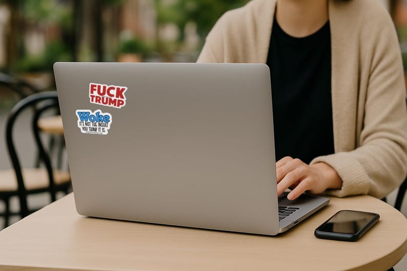 May include: A silver laptop with two stickers on the lid. The top sticker is red with white text reading FUCK TRUMP. The bottom sticker is blue and white with text reading Woke IT'S NOT THE INSULT YOU THINK IT IS. The laptop is open on a table.