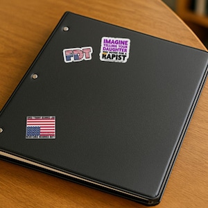 May include: A black three-ring binder featuring three stickers. One sticker reads FDT in a red, white, and blue pattern. Another reads IMAGINE TELLING YOUR DAUGHTER YOU VOTED FOR A RAPIST. The third is a flag with text WHEN TYRANNY BECOMES LAW.