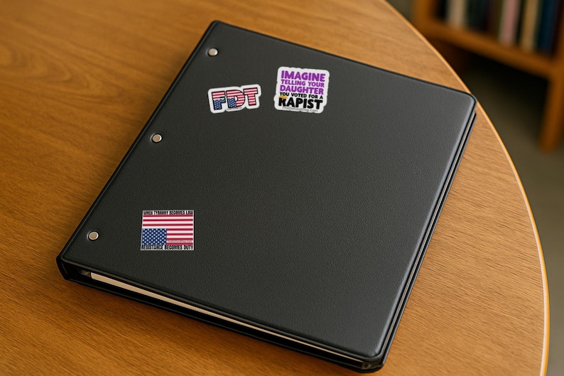May include: A black three-ring binder featuring three stickers. One sticker reads FDT in a red, white, and blue pattern. Another reads IMAGINE TELLING YOUR DAUGHTER YOU VOTED FOR A RAPIST. The third is a flag with text WHEN TYRANNY BECOMES LAW.