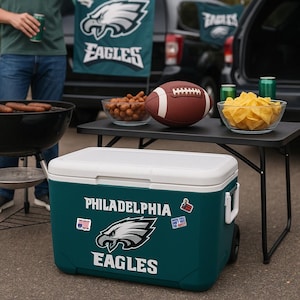 May include: A teal rolling cooler with a white lid featuring the Philadelphia Eagles logo and text. The cooler is decorated with small stickers, including an American flag, and sits on the ground in front of a table with a football and snacks.