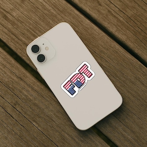 May include: A light-coloured smartphone with a dual-lens camera system on the back. A sticker is applied to the centre of the phone, featuring the letters FDT in a bold, block font. The letters are filled with a red, white, and blue American flag pattern.