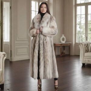 May include: A long, light-coloured fur coat with a large collar. The coat is worn by a person standing in a well-lit room with hardwood floors. The coat appears to be made of soft, luxurious fur.