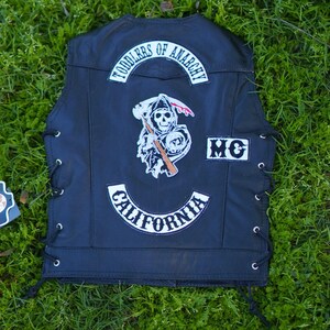 Puede incluir: Chaleco de cuero negro con parches blancos. La parte trasera del chaleco presenta un gráfico de la Parca, las palabras "TODDLERS OF ANARCHY" y "CALIFORNIA". El chaleco tiene cordones laterales y las letras "MC".