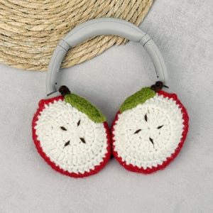May include: Gray headphones with crocheted apple-shaped ear covers. The ear covers are white with red edges, green leaves, and brown stems. The headphones rest on a woven, circular mat.