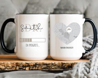 May include: White ceramic mug with a black handle and rim. One side features the name "Smith" with "DR." above and "IN PROGRESS..." below a progress bar. The other side has a heart-shaped map of Harvard University.