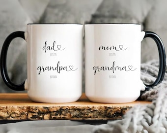 May include: Two white ceramic mugs with black handles and rims. One mug reads "dad grandpa" with "est 1995" and "est 2020". The other mug reads "mom grandma" with the same dates. The mugs are on a wooden surface.