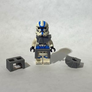 Peut inclure: Une figurine Lego d'un clone trooper blanc et bleu avec une armure grise. Deux accessoires Lego gris sont de chaque côté de la figurine. La figurine a une bande bleue sur le casque et des accents bleus sur l'armure.
