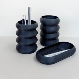 Può includere: Un set di tre organizer da scrivania blu scuro. Un portapenne alto e scanalato contiene penne e un righello. Due contenitori aggiuntivi sono impilati e un vassoio ovale contiene penne. Gli oggetti sono su una superficie chiara.