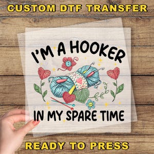 May include: A DTF transfer with the text "I'M A HOOKER IN MY SPARE TIME" in black, featuring yarn, crochet hooks, and hearts. The design includes the text "CUSTOM DTF TRANSFER" and "READY TO PRESS" in yellow.