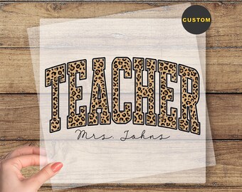 May include: A custom DTF transfer with the word "TEACHER" in a leopard print design. The name "Mrs. Johns" is below the word. The words "CUSTOM" and "READY TO PRESS" are also visible.