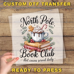 May include: A DTF transfer featuring a whimsical design for a book club. The design includes a mug of hot cocoa topped with whipped cream, a gingerbread man, and a candy cane. The words "North Pole Book Club" and "Hot cocoa served daily" are also included.