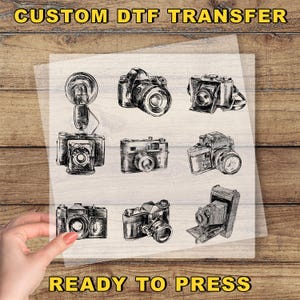 May include: A sheet with a collection of vintage camera illustrations in black and white. The text "CUSTOM DTF TRANSFER" is at the top, and "READY TO PRESS" is at the bottom.