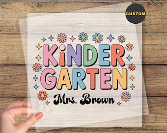 May include: A colorful DTF transfer with the words "KINDER GARDEN Mrs. Brown" in a retro font, surrounded by flower and star graphics. The text "READY TO PRESS" is at the bottom. The transfer is held by a hand.