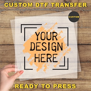 May include: A custom DTF transfer with the text "YOUR DESIGN HERE" in black, set against an orange splash design. The words "CUSTOM DTF TRANSFER" and "READY TO PRESS" are also visible. The transfer is held by a hand.