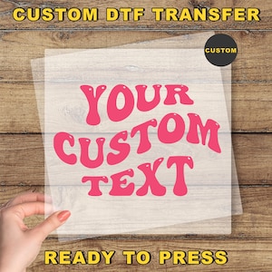 May include: A clear transfer sheet with the words "YOUR CUSTOM TEXT" in pink, retro-style font. The words "CUSTOM DTF TRANSFER" and "READY TO PRESS" are in yellow. A black circle with the word "CUSTOM" is in the upper right corner.
