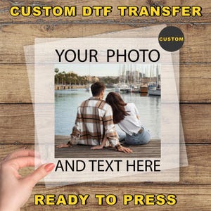 May include: A clear DTF transfer sheet with the text "CUSTOM DTF TRANSFER", "YOUR PHOTO", "AND TEXT HERE", and "READY TO PRESS". The transfer features a photo of a couple sitting by the water. A hand is holding the transfer.