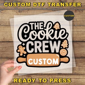May include: A DTF transfer with the text "The Cookie Crew" in black and white, featuring gingerbread men, Christmas trees, and a rolling pin. The word "Custom" is also included. The transfer is ready to press.